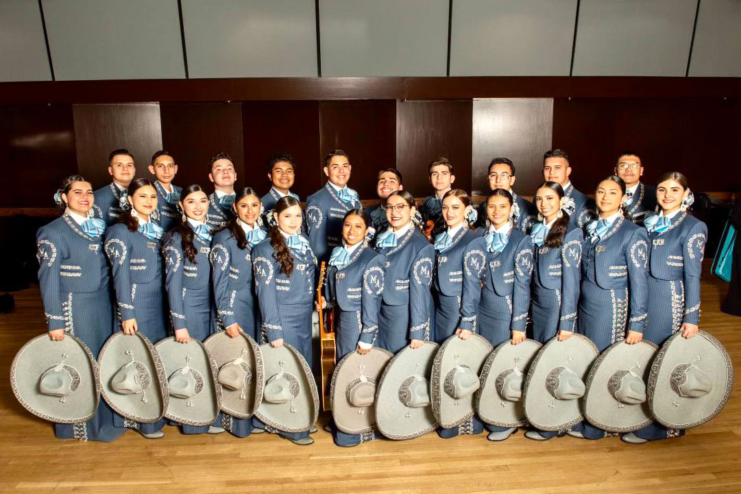 Twenty-two students in matching blue suits with large brimmed hats in hand pose in a classroom.