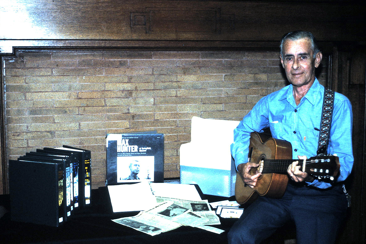An elder white man sits on a table holding an acoustic guitar. On the table are newspaper clippings and black binders.