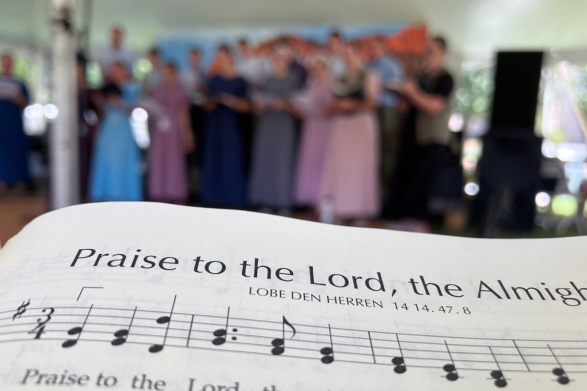 In the foreground, close-up on a songbook opened to sheet music with the song title Praise to the Lord, the Almighty. In the background, out of focus, a choir performs under a tent.