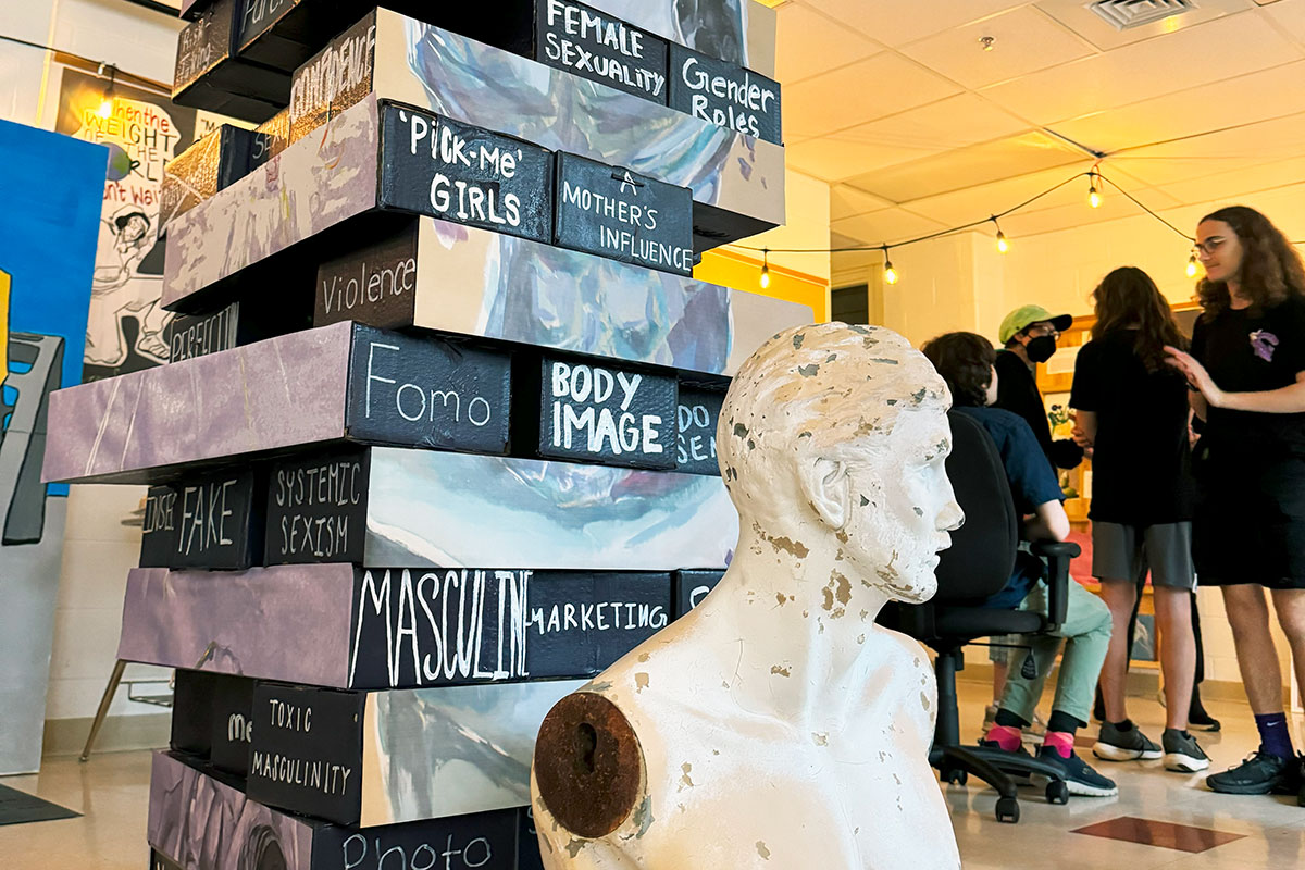 A pop-up museum display, featuring a tower of several “bricks” stacked like Jenga pieces. Each brick has writing on it: Fomo, body image, masculine marketing, pick-me girls, female sexuality, gender roles, a mother’s influence. In front of the tower is a white, armless bust sculpture, and behind it is a small crowd of teens chatting.