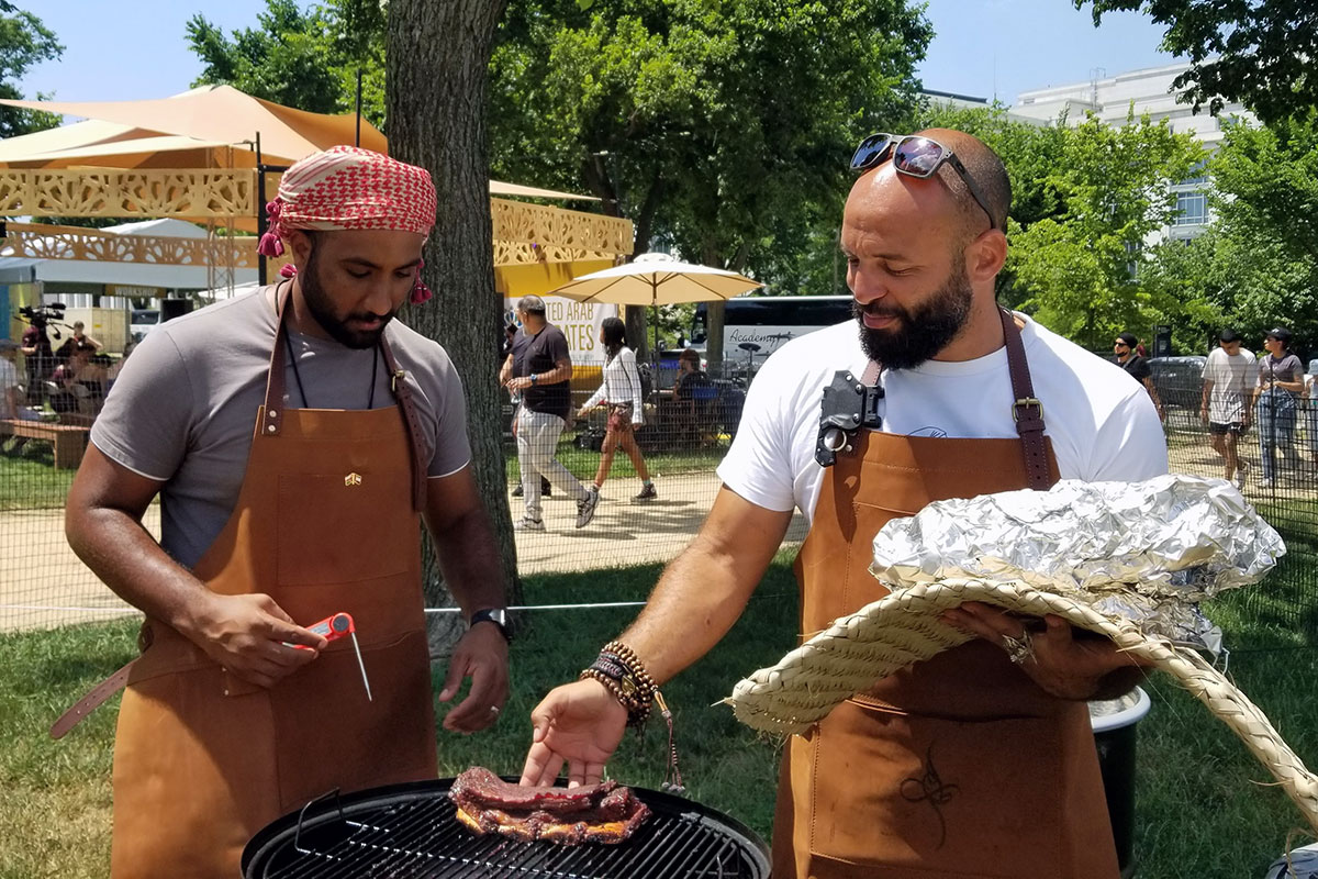 Two men, both wearing tan leather aprons, tend to a piece of meat on an outdoor grill. One is holding a meat thermometer, and the other a tinfoil-wrapped bundle and a woven mat.
