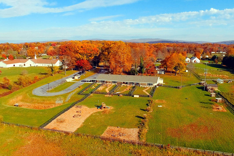 Aerial shot of a single-story building and farmland during the fall, with green pastures and trees in the distance with red and orange foliage. 