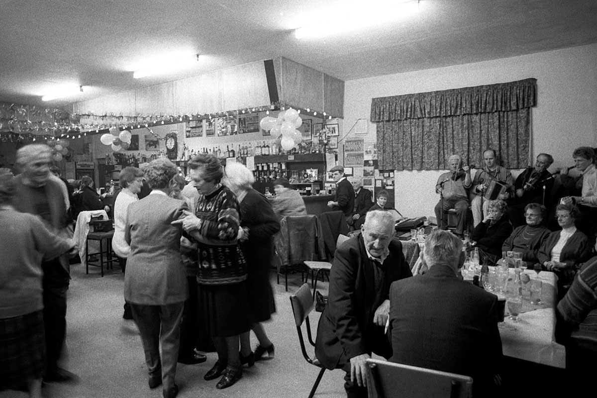 Couples dance and sit at tables in a bar, with a few musicians lined up along one wall. Black-and-white photo.