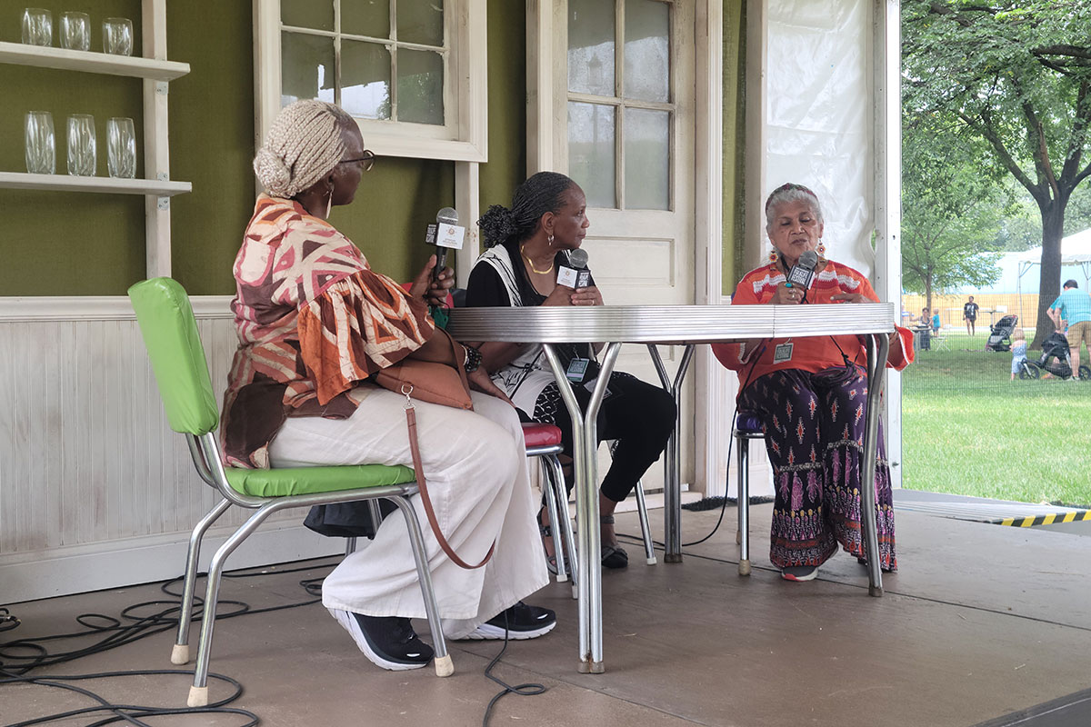 Three women hold microphones, sitting around a Formica table on an outdoor stage.