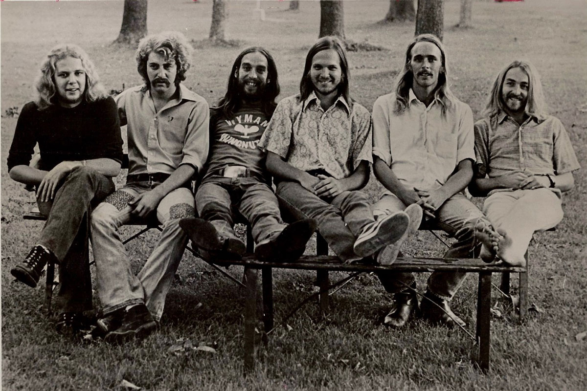 Six young men, most with long hair, pose sitting on a picnic bench outside. Black-and-white photo.