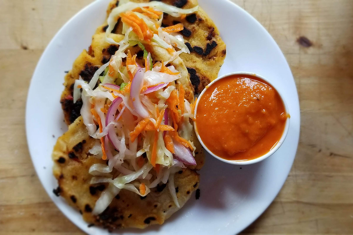 Close-up on a plate of fried pupusas, with shredded pickled vegetables on top and a small dish of red sauce on the side.