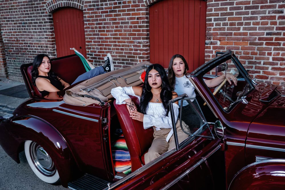 Three women with long dark hair pose in a shiny, port wine-colored classic convertible car with the top down.