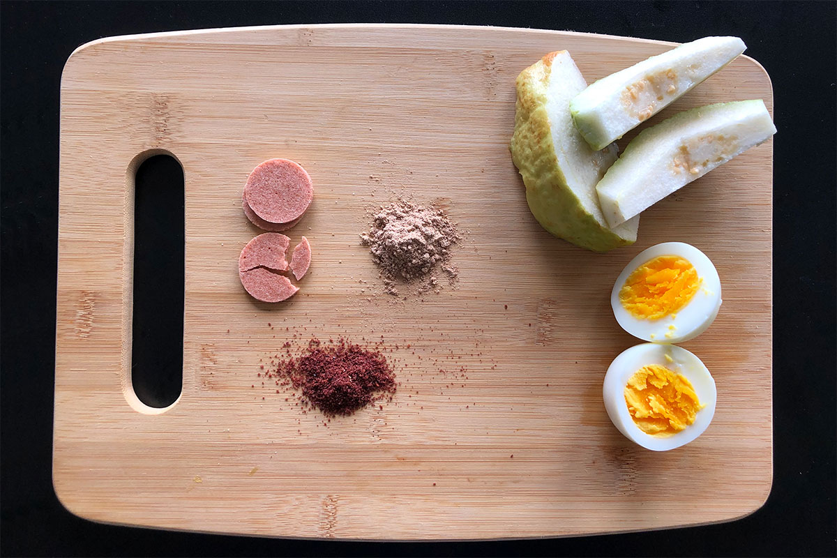 Cutting  board containing a lineup of snacks: pink circles, green and white slices of guava, a hard-boiled egg, and two pinched of pinkish powders.