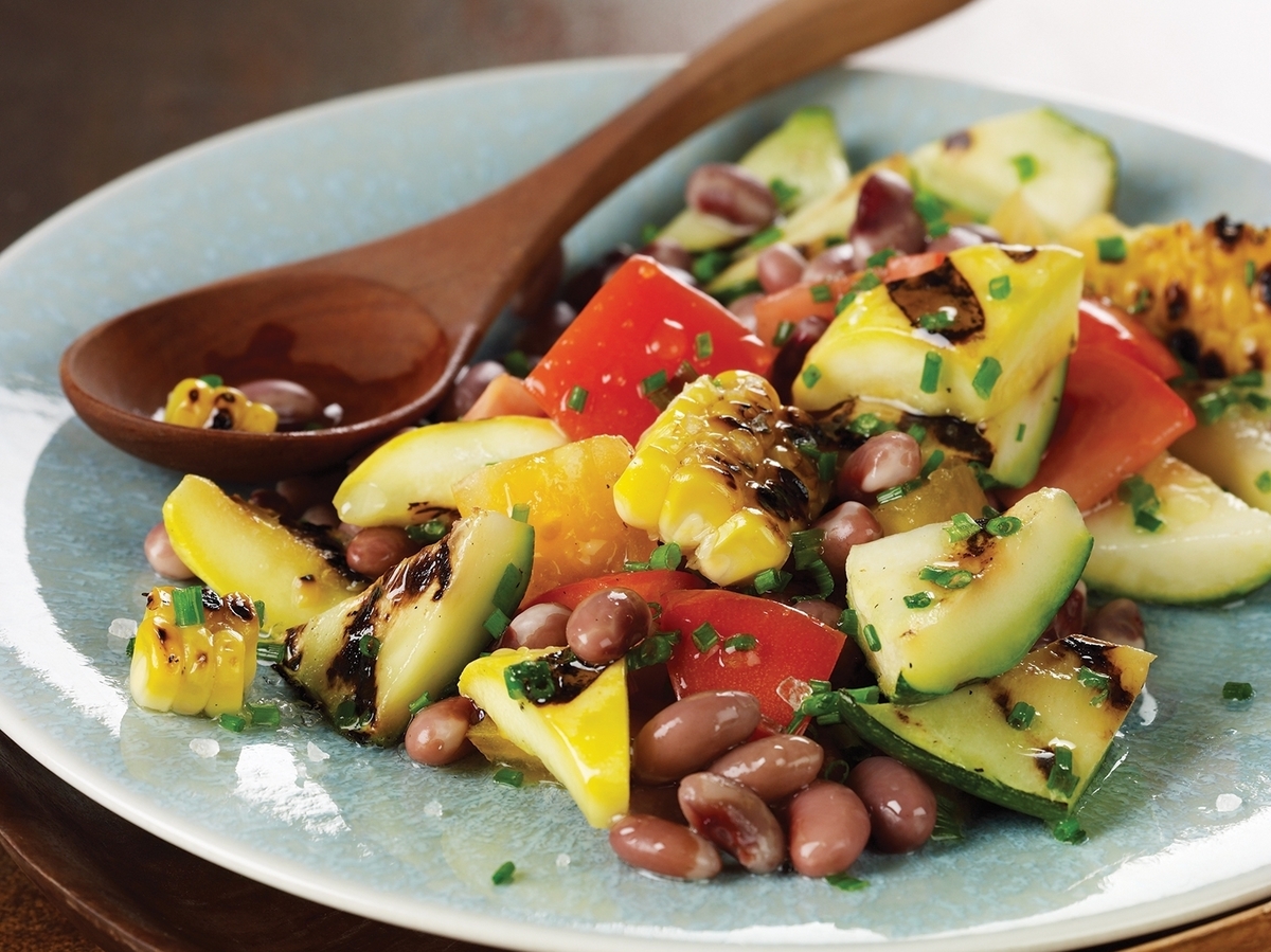 Photo of a plate full of cooked yellow corn, reddish beans, green zucchini, yellow squash, and red tomatoes, with a carved wooden spoon sticking out.