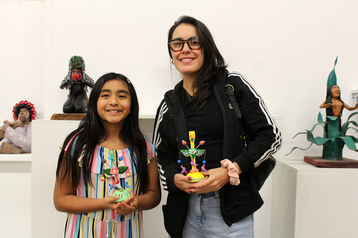A young woman and young girl hold up painted, decorated tree sculptures approximately six inches tall, both smiling for the camera. Behind them, more sculptures against white gallery walls.