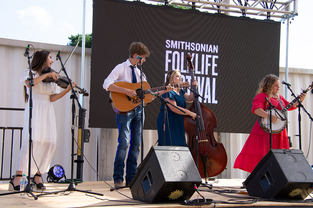 Four teens performing on stage, each on a string instrument: fiddle, guitar, upright bass, and banjo. A screen behind them shows black and white Smithsonian Folklife Festival logo.