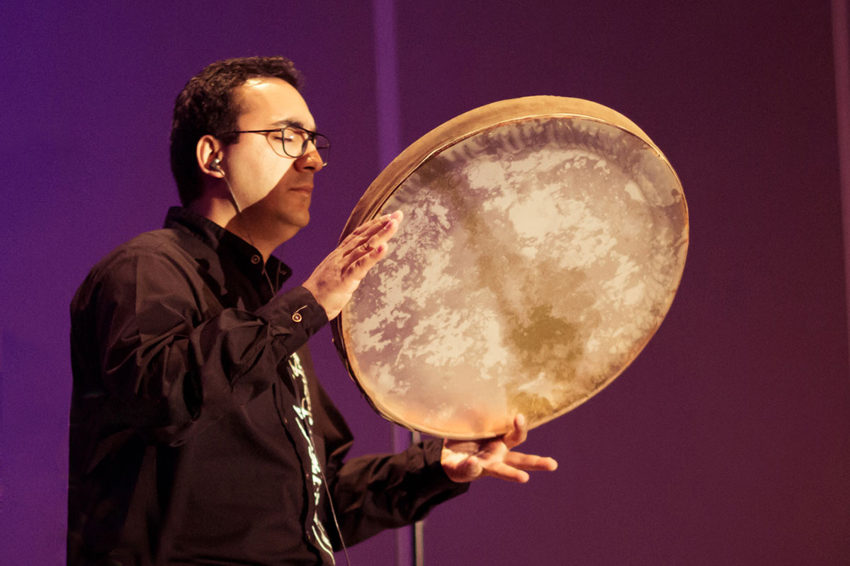 A man plays a large frame drum, held in one hand and struck with the other.