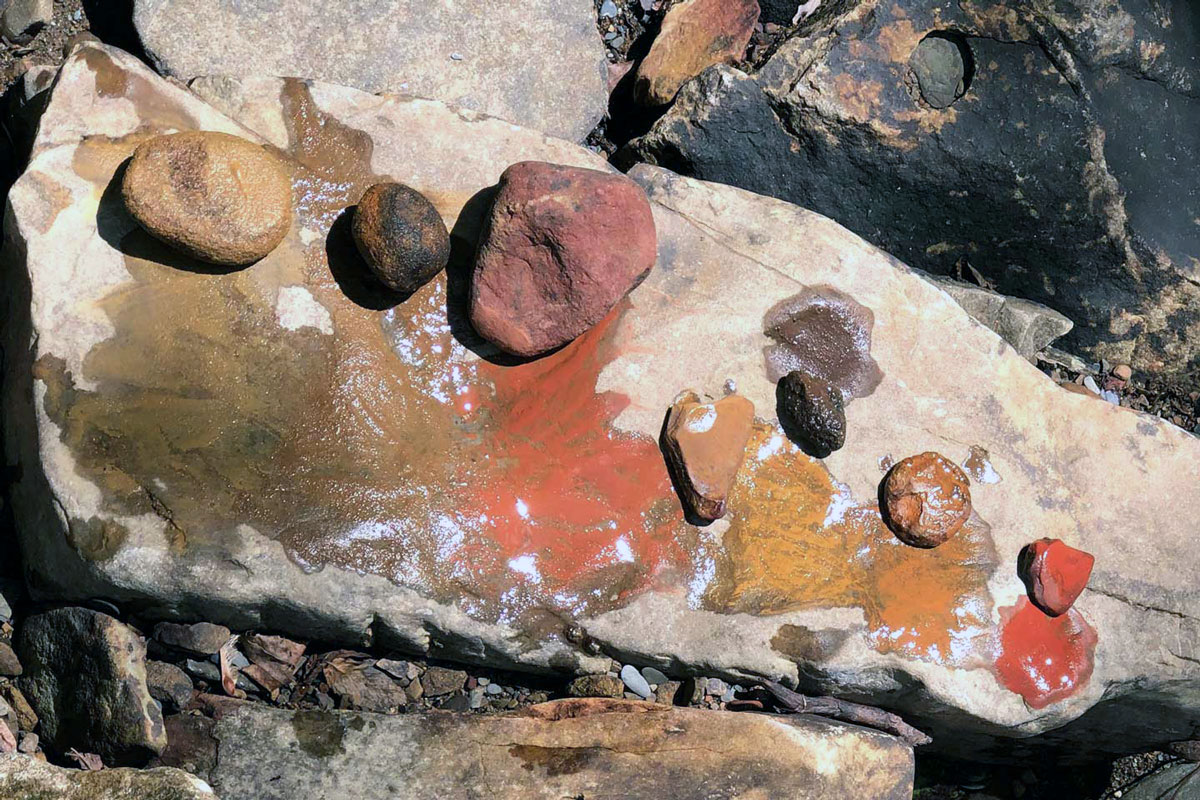 Seven small stones, arranged in a line on a larger flat stone, have each produced a small, wet puddle of colored pigment, in shades of brown, red, and orange.