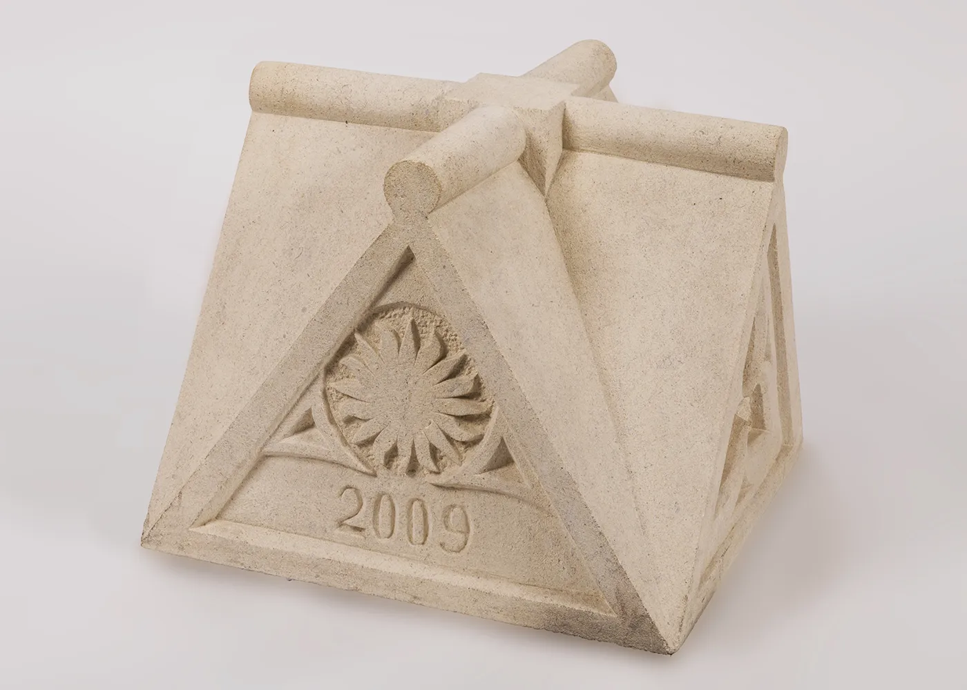 A block of carved white limestone with four three-dimensional triangular sides, one with a carving of a multi-pointed sunburst and the year “2009.”