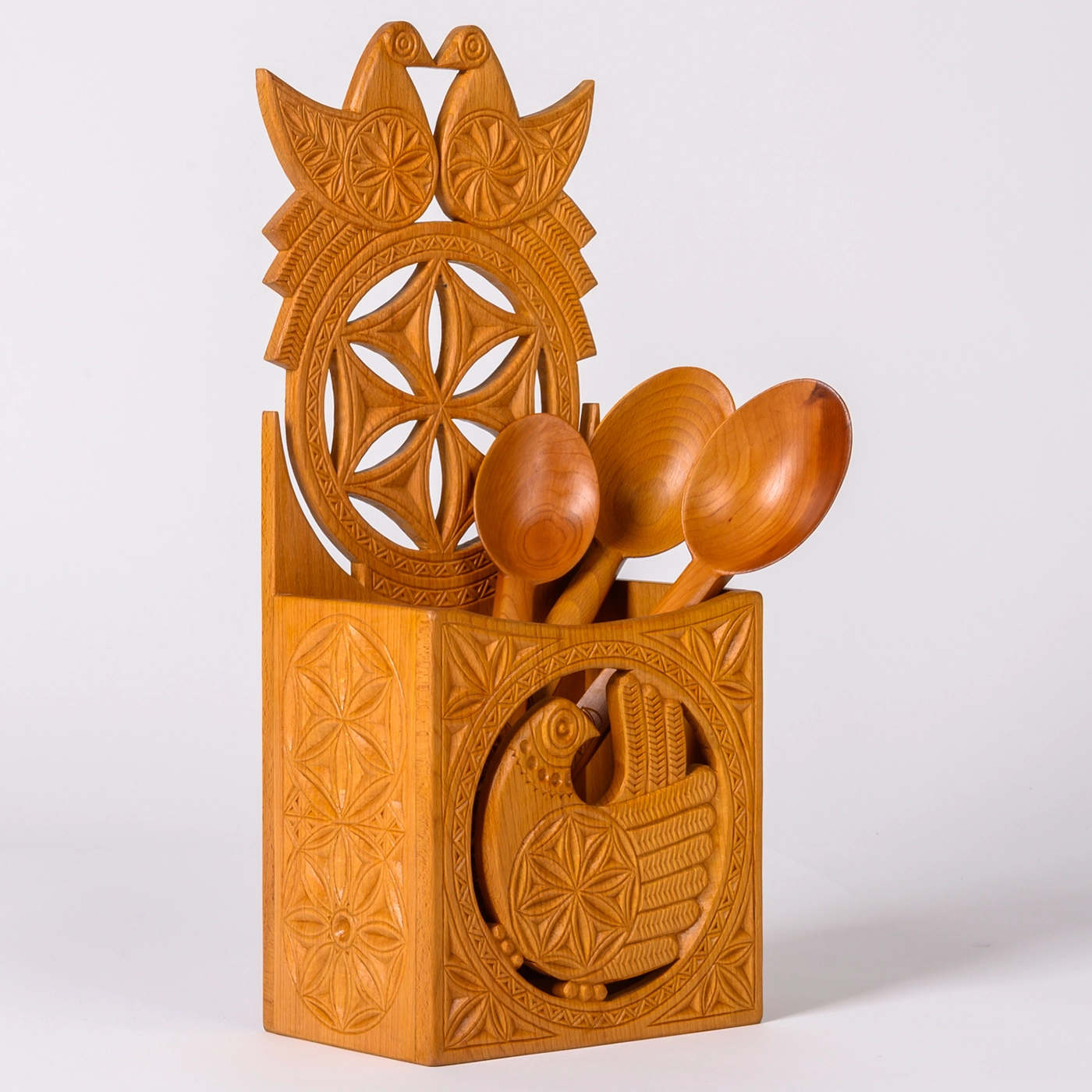 A carved wood kitchen storage container decorated with surface and cut-out geometric, plant, and bird designs holds three carved wooden serving spoons standing upright. 