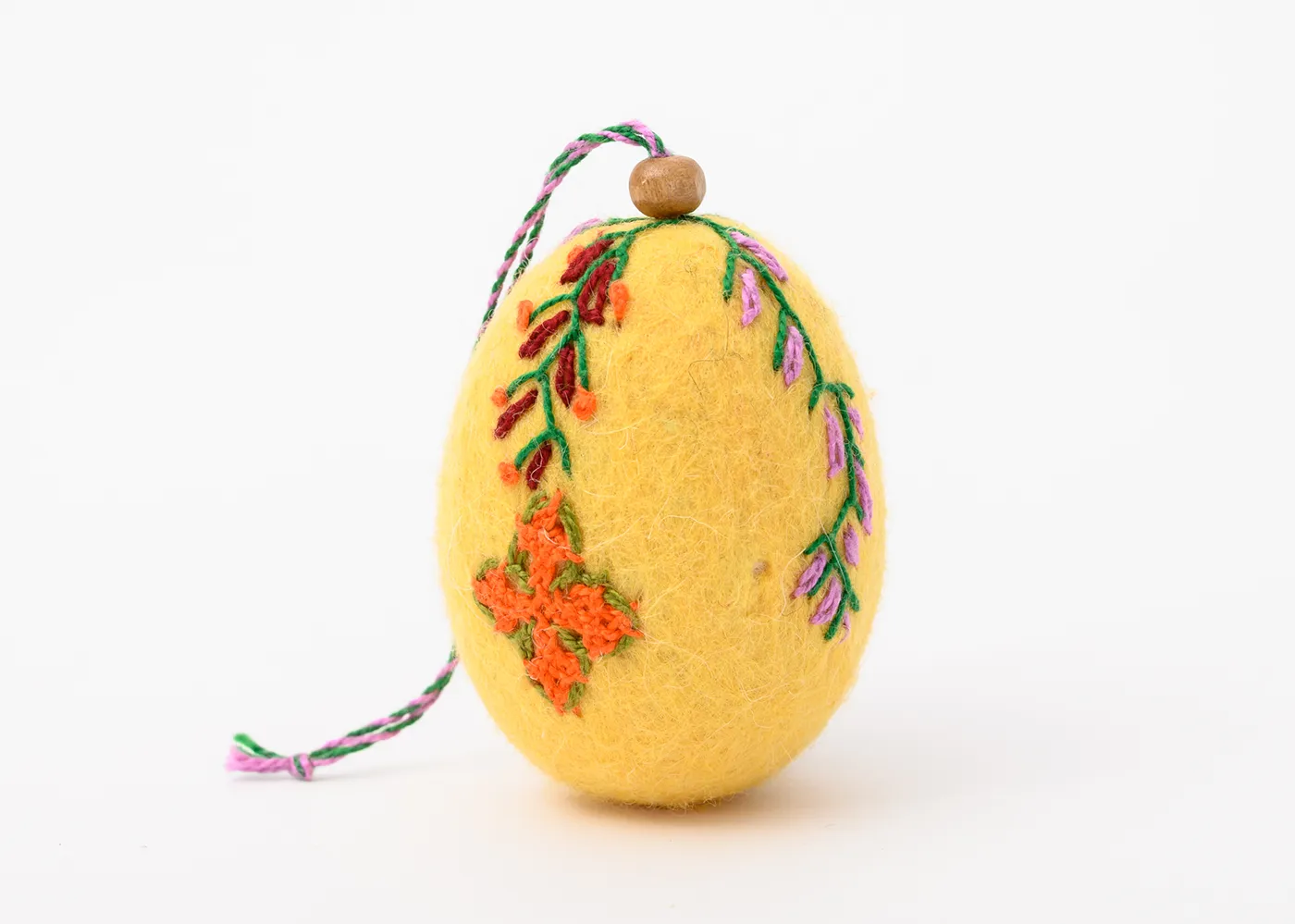 An egg-shaped ornament made of yellow felt is decorated by two colorful strands of floral embroidery that extend midway down the length of the egg. The one on the left terminates in an orange cross.