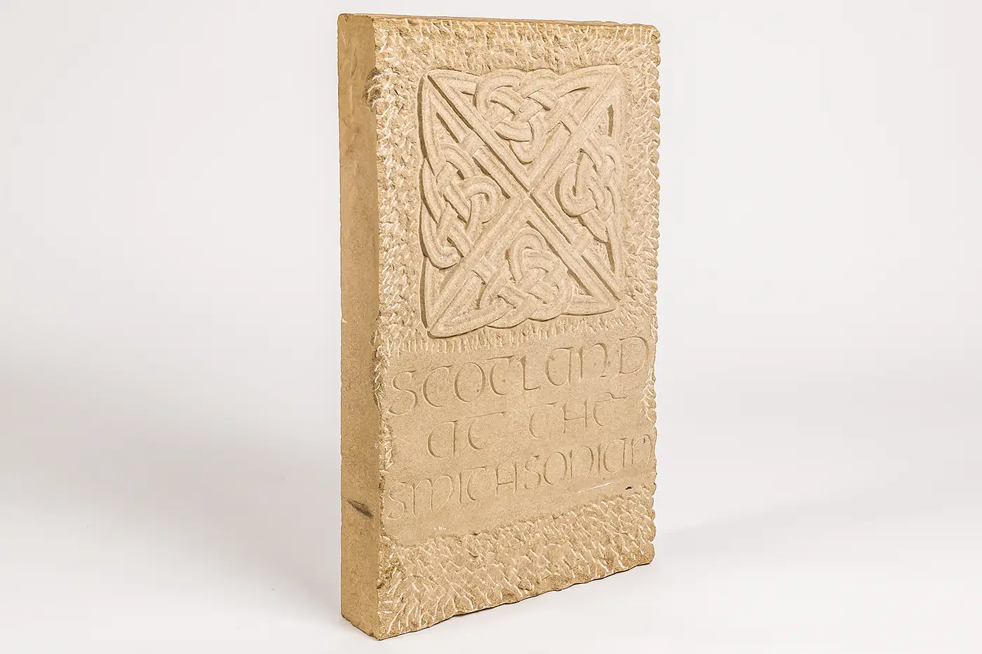 An upright, rectangular slab of thick sandstone with a carved Celtic knot at the top, above the words “Scotland at the Smithsonian” carved in the lower half. 