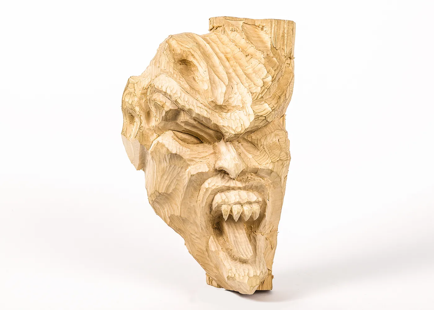 A partially carved, unpainted wood mask depicts a deeply chiseled, grimacing devil’s face with furrowed brows, a creased forehead, and open mouth with sharply pointed upper teeth.  