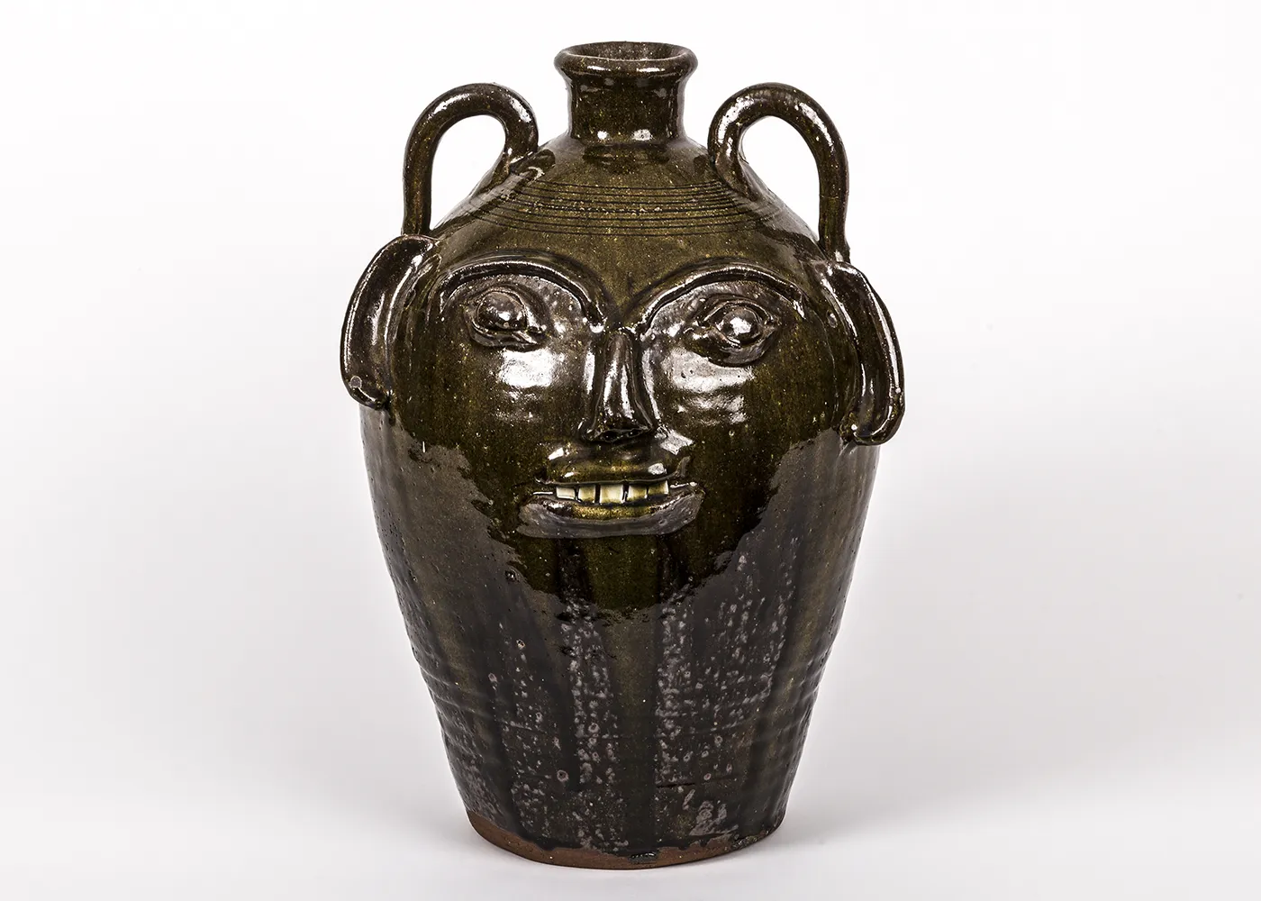 A tall, dark brown ceramic “face jug” with a glossy glaze includes two looped handles set high on the shoulders, below which are facial features that include ears, eyes, nose, and mouth.  
