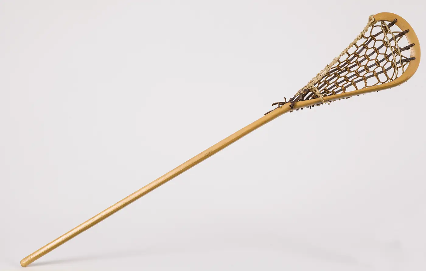 A natural, light colored wooden lacrosse stick, featuring a basket woven with strips of rawhide. 
