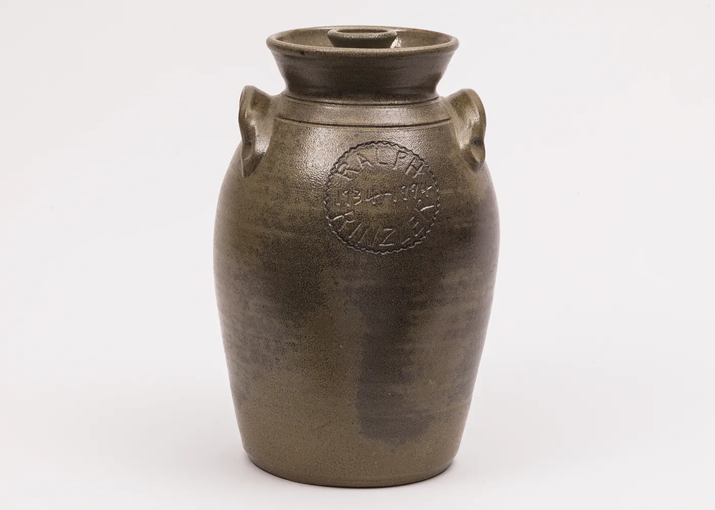 A clay pot, wider at the top and tapering to the base, covered in a dark brown glaze, and incised with the name “Ralph Rinzler” in all caps and the dates “1934–1994.” 
