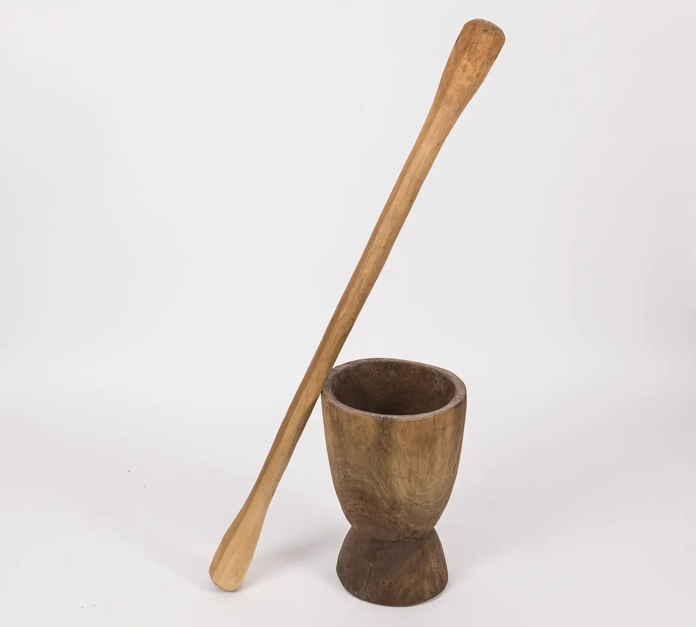 A tall, carved, light-colored wooden pestle with two bulbous ends, rests at an angle against a large, rounded, carved mortar vessel of a darker wood grain. 