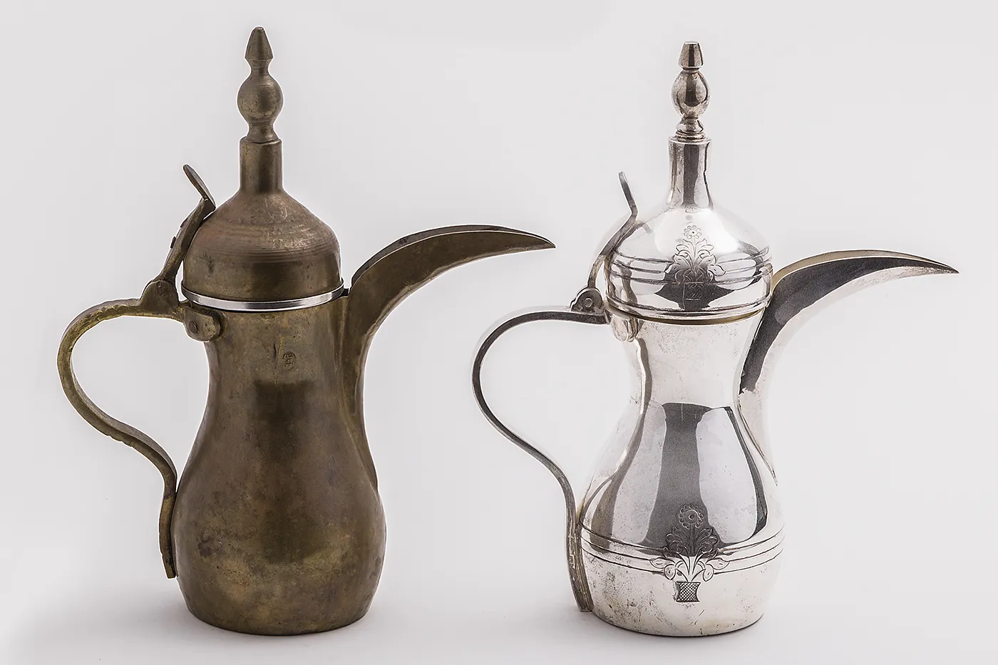 Two tall, narrow coffee pots—one copper and one silver—with deep, wide spouts; large, curved handles; and tall cylindrical lids.