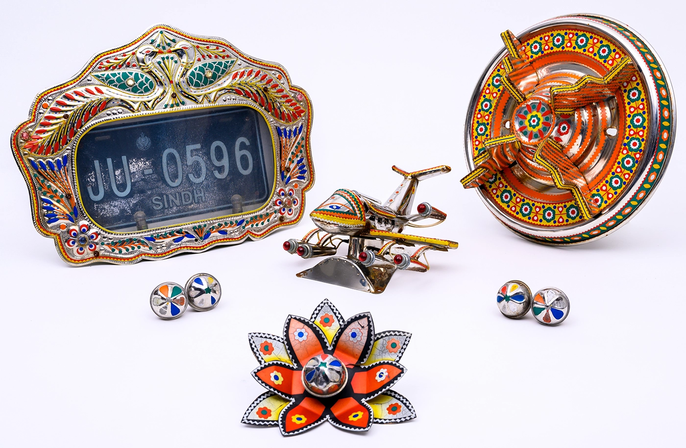 A collection of six colorfully decorated chrome accessories from the exterior of a Pakistani cargo truck: a license plate holder, hood ornament, hubcap decoration, ornamental flower and bells—all decorated with colorful vinyl tape.