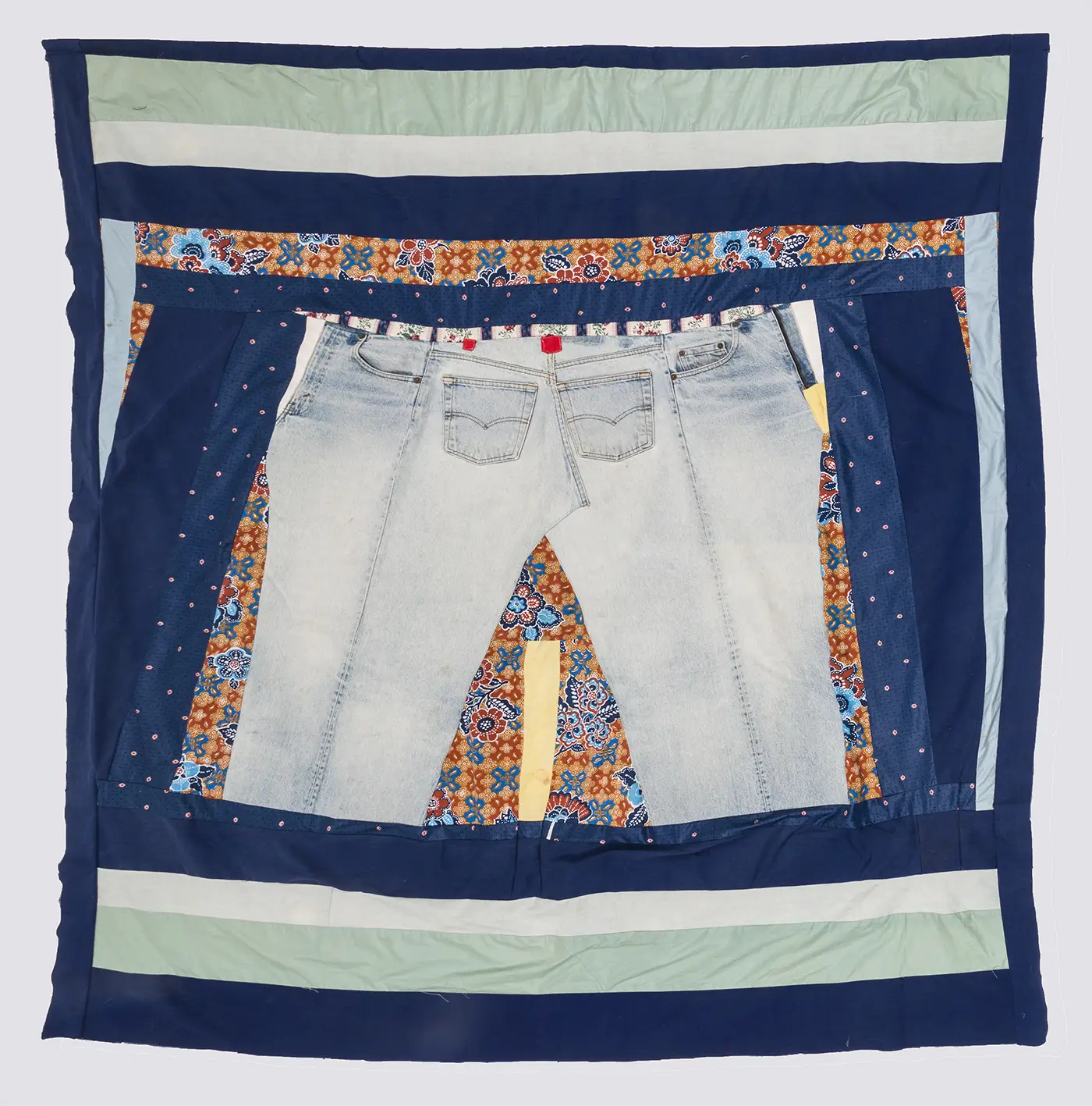 A patchwork quilt with navy blue border framing a pair of faded blue jeans at the center, surrounded by a variety of fabric scraps of different sizes and shapes in orange, blue, seafoam green, and white. 