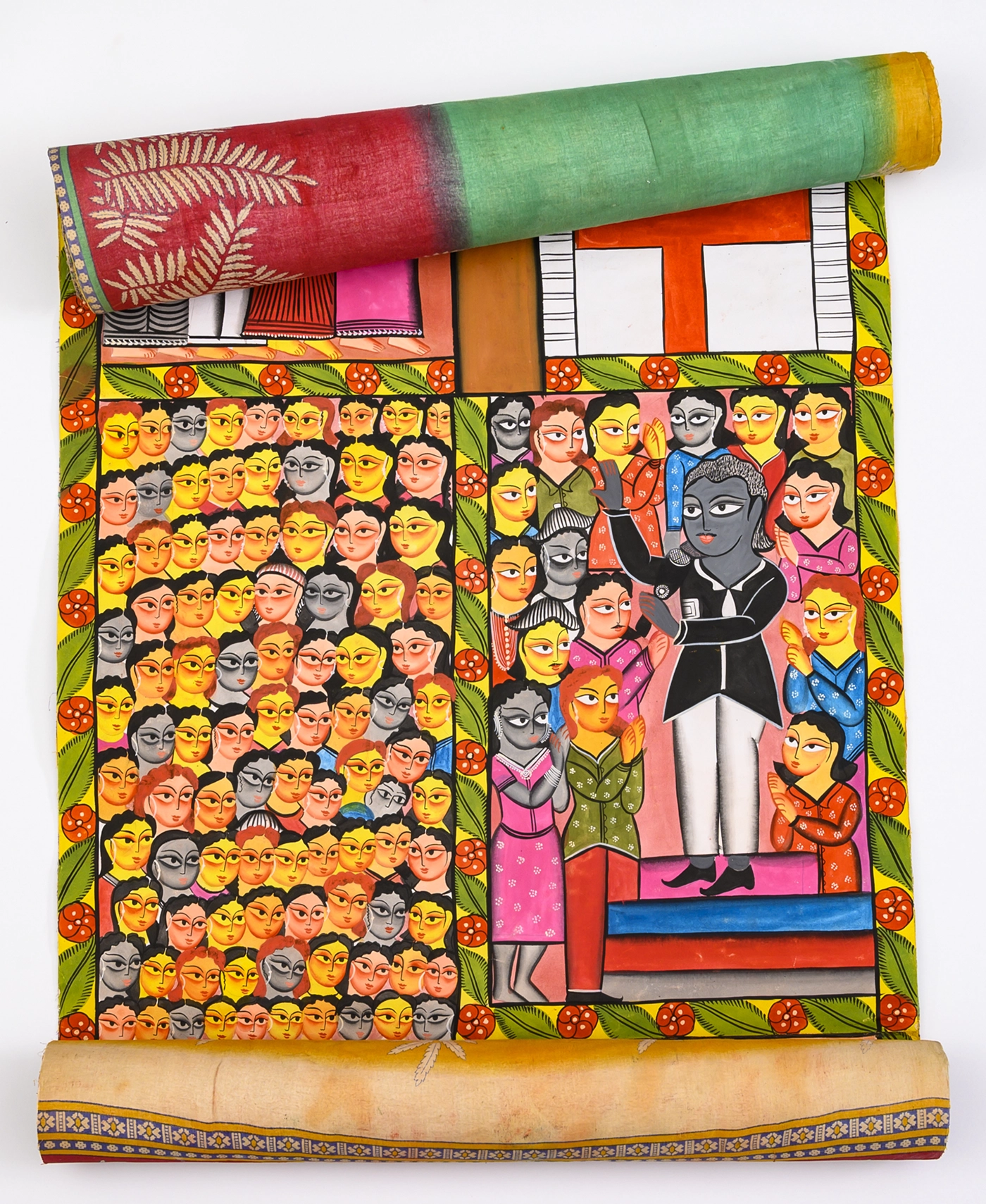 A partially unrolled Indian “story scroll” painting with a multicolored fabric backing features two side-by-side scenes. On the left is a closely packed crowd of faces. On the right, a man holds a microphone and speaks to a close-in group of people surrounding him. 