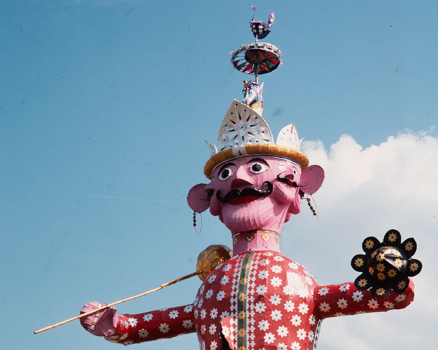 A giant papier-mâché, multi-faced, mustached figure in shades of pink, red, and white is seen outside against a sunny, bright blue sky, and tethered to the ground with guide wires. 