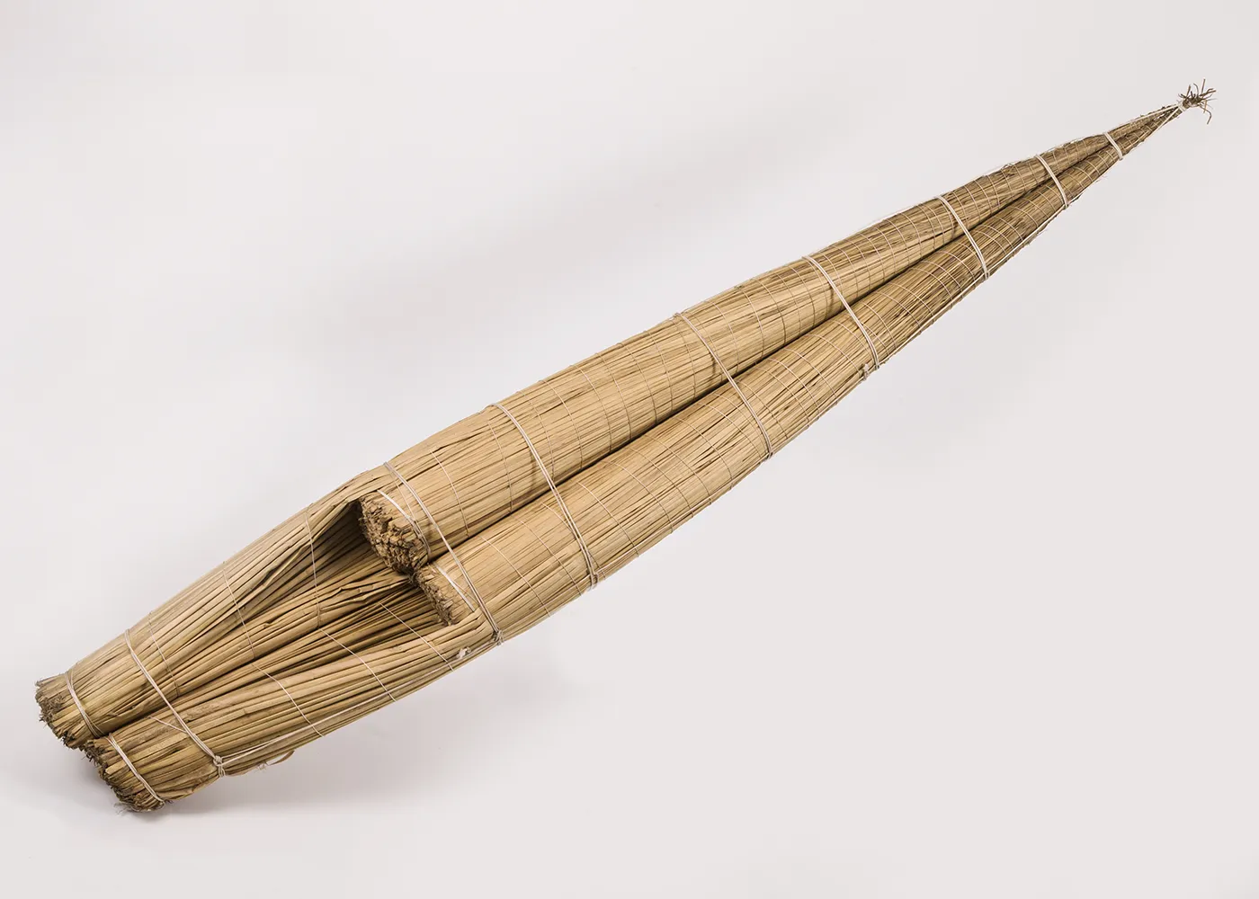 A narrow kayak-shaped raft made of light-colored reeds tightly packed and bound together with twine. From the blunt back end, the raft narrows to a pointed tip in the front, which curves slightly upward. 