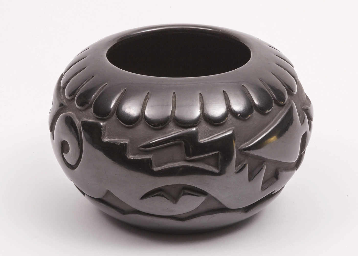 A black, rounded, highly polished bowl with a wide opening at the top and deeply carved relief designs which encircle the vessel. 