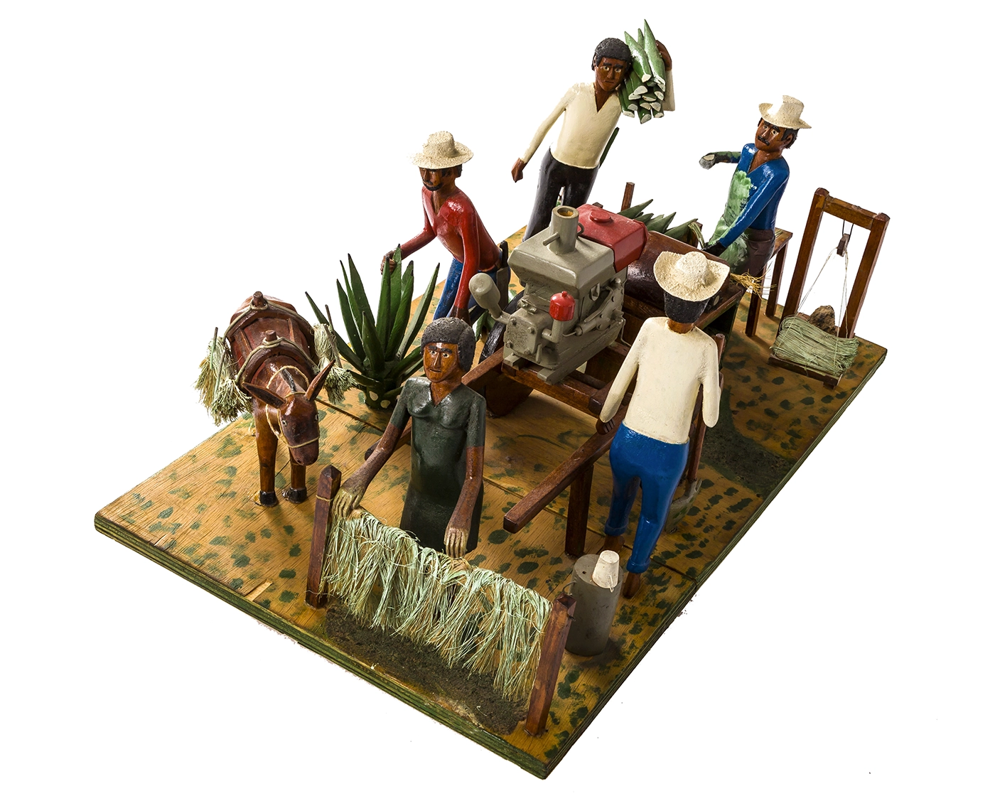 A tableau of hand-carved and painted wood figures engaged in work activities related to making rope is arranged on a painted plywood surface. Four men, one woman, one donkey, a machine, and racks holding the fiber are included.  
