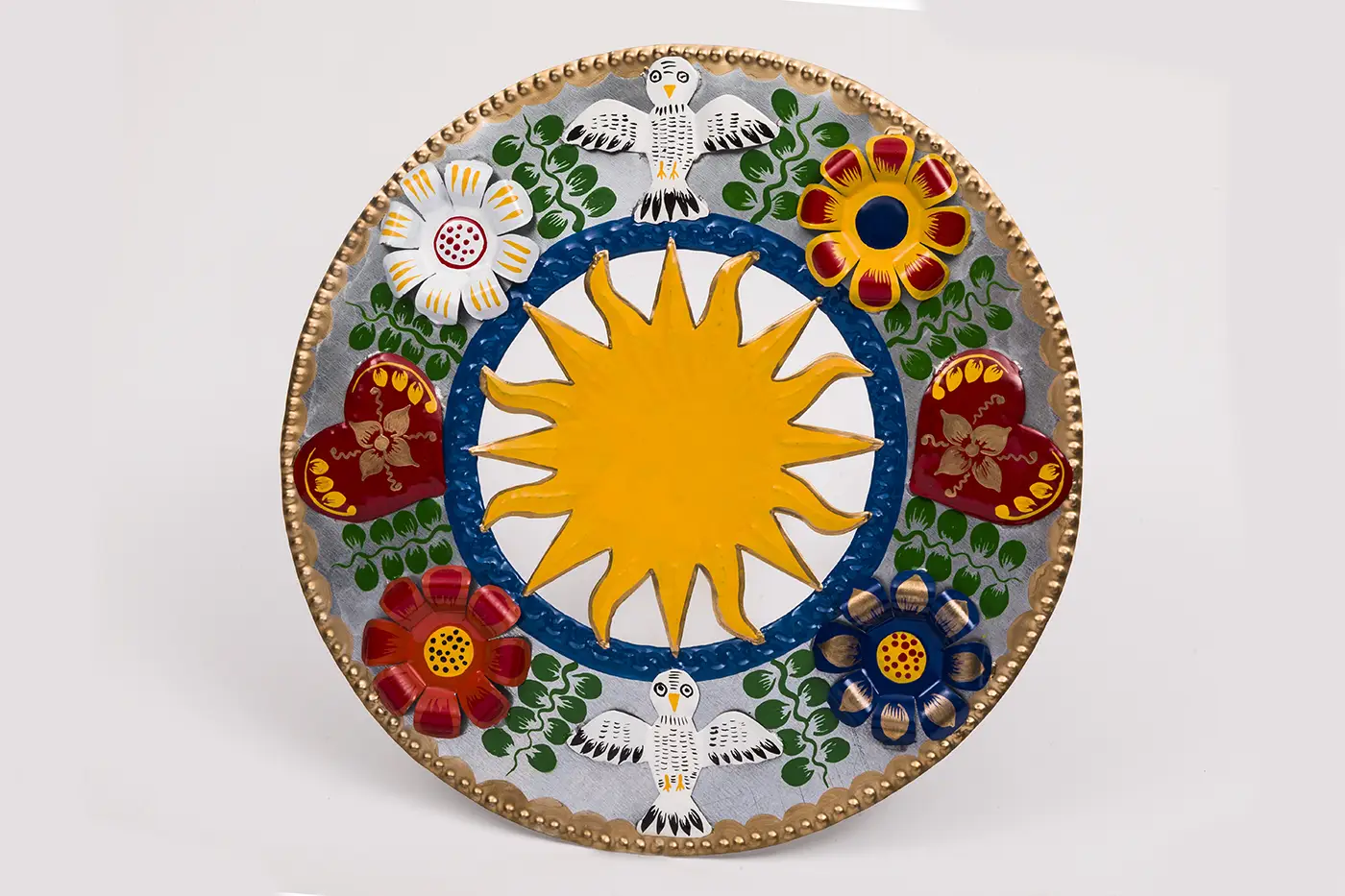 A colorful round plaque made of cut, shaped, and painted tin decorated with red, white, and blue flowers attached to the rim, two white birds at top and bottom, and a large yellow, multi-pointed sunburst in the center. 