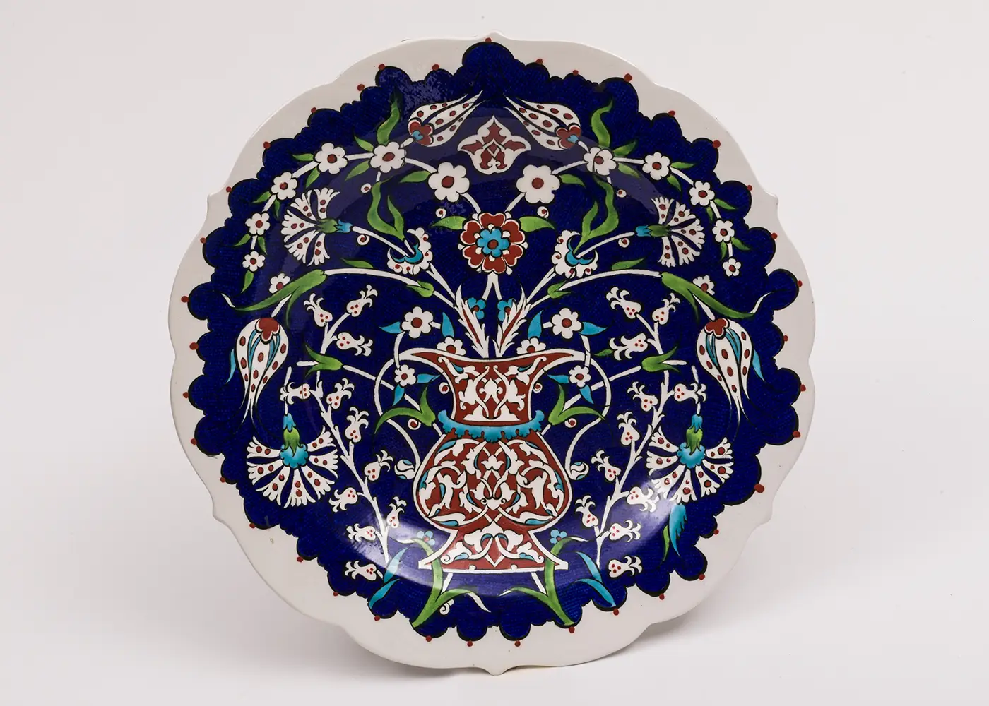 A white porcelain plate with scalloped edges is filled with a painted delicate design of flowers arching from a vase against a dark blue background. 