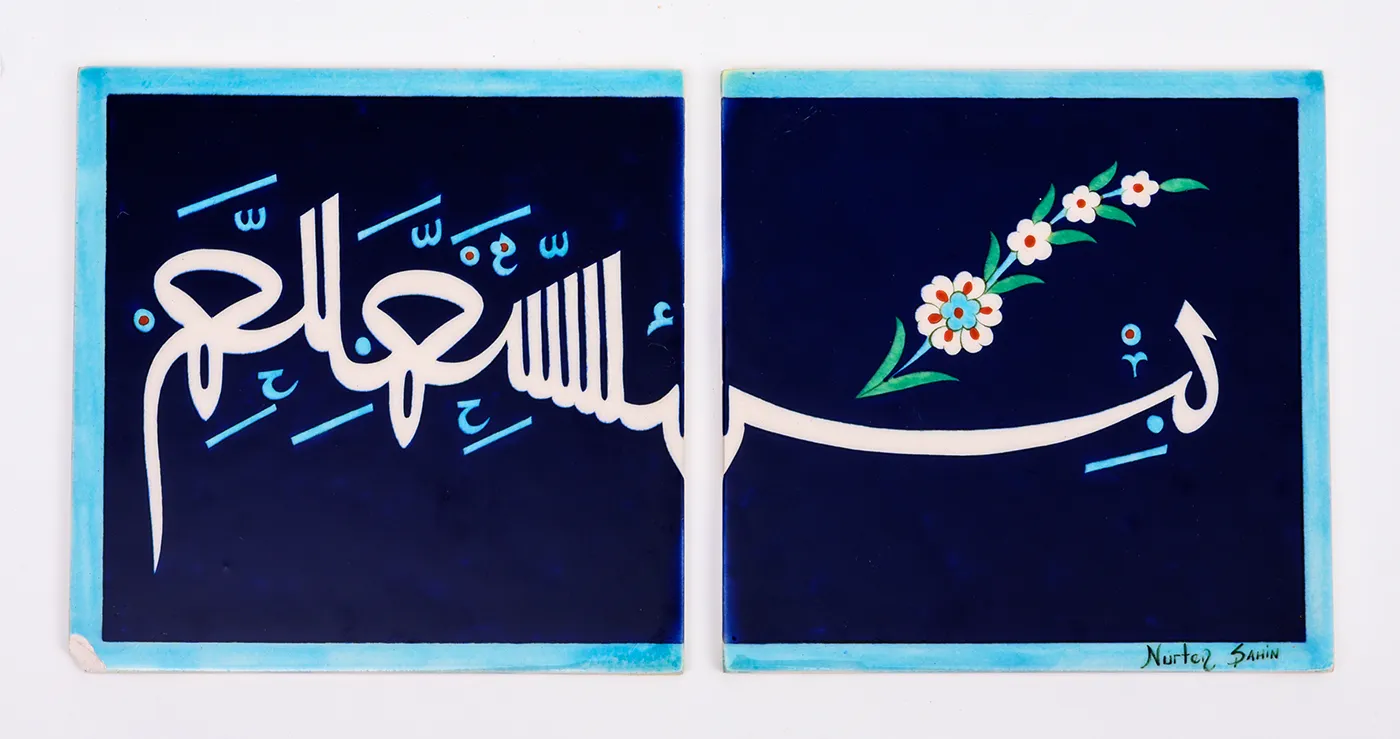 Two square ceramic tiles decorated with a design of white Arabic calligraphy and a spray of white flowers that extends across both tiles, set off against a navy-blue background with light blue border. 