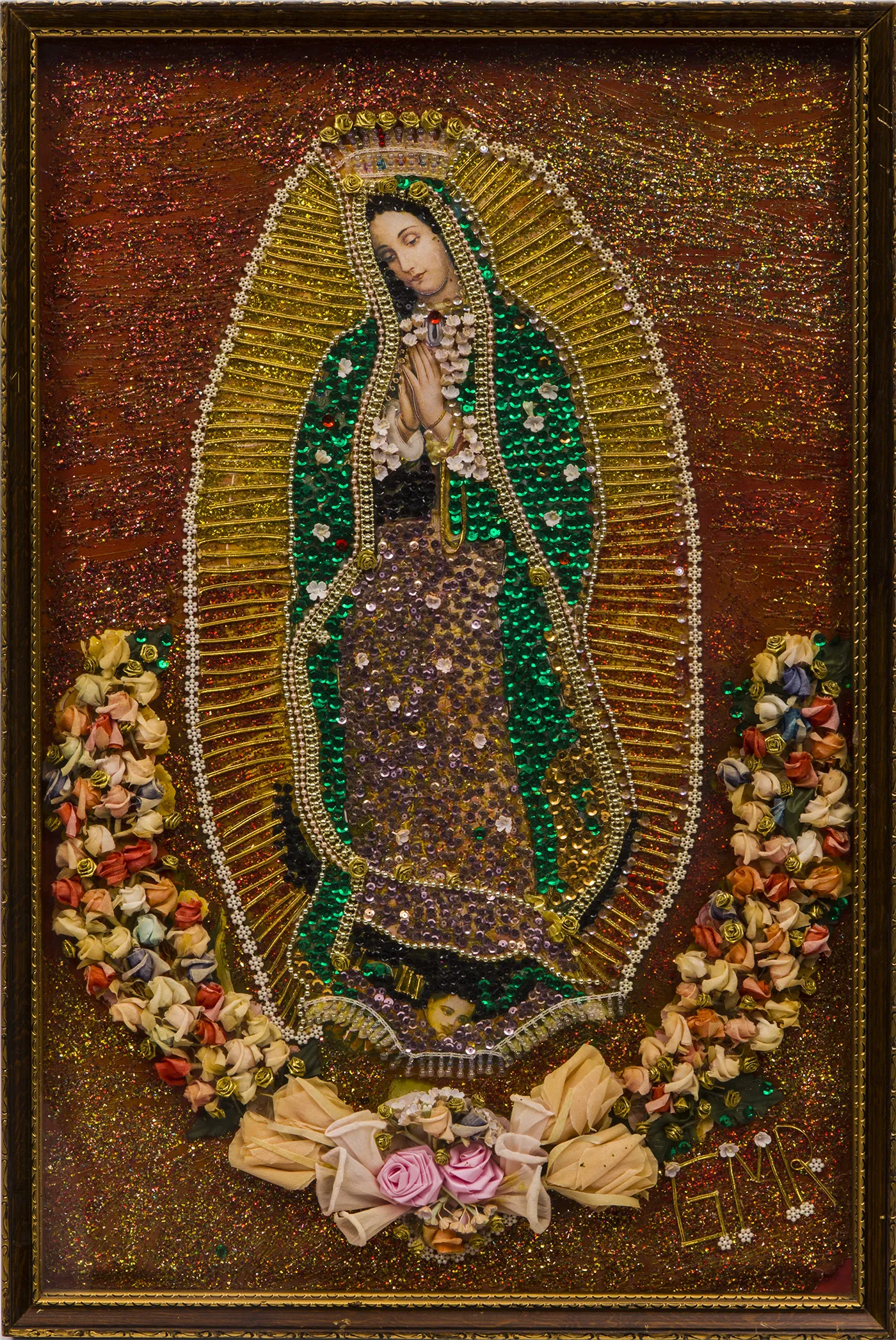 A framed, mixed-media composition of the Virgin of Guadalupe, featuring sparkling beading in shades of green, gold, maroon, and white and framed at the bottom with an open semi-circle of delicate paper roses.   