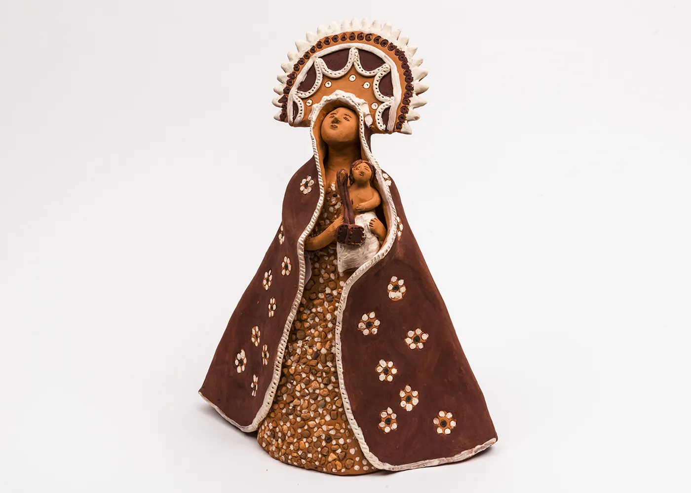 A clay figure of the Virgin Mary holding baby Jesus features colors of red, orange, and white, and is textured with natural pebbles. 