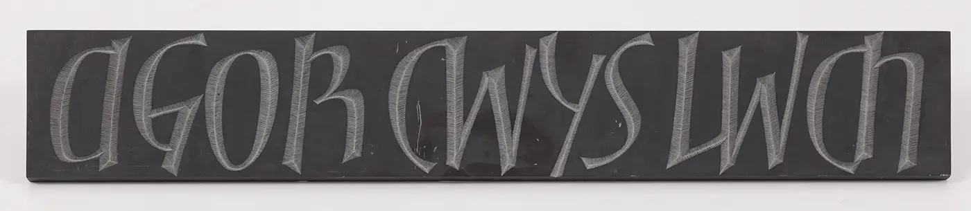 A long rectangular slab of black slate carved with evenly spaced, uppercase, rounded letters that appear light gray and read, in Welsh, “Agor Cyws lwch.” 