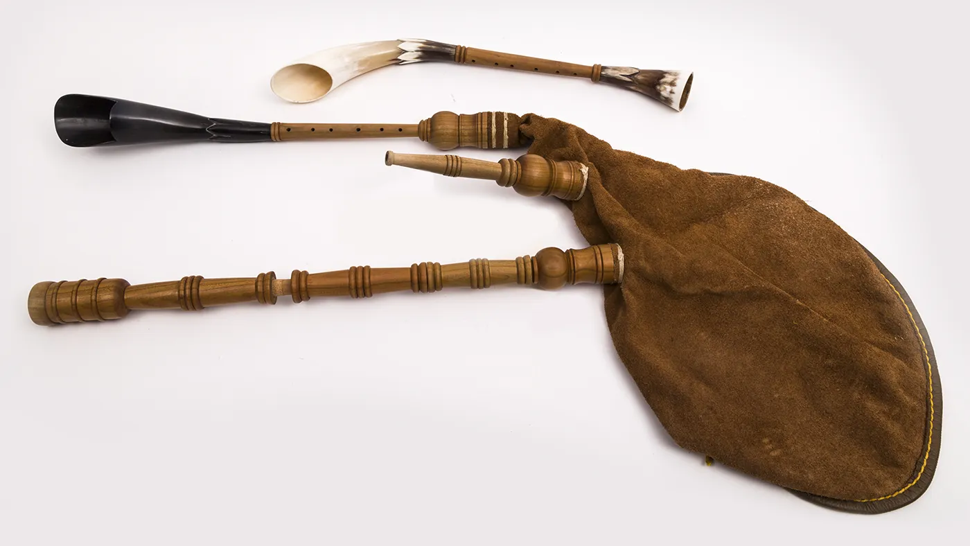 A hornpipe made of carved horn and turned wood sits next to a bagpipe with a brown leather pouch and pipes of carved horn and turned wood. 