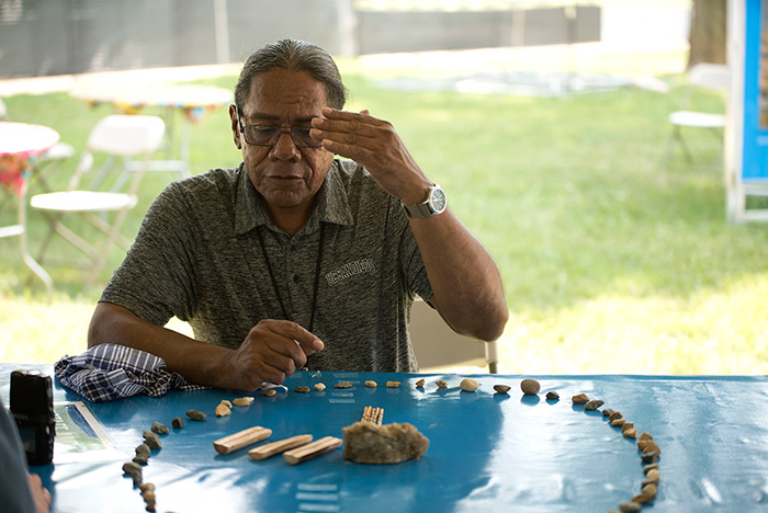 A man sits at an outdoor table, holding up one hand, presiding over a game using sticks and stones laid out in a circle in front of him.