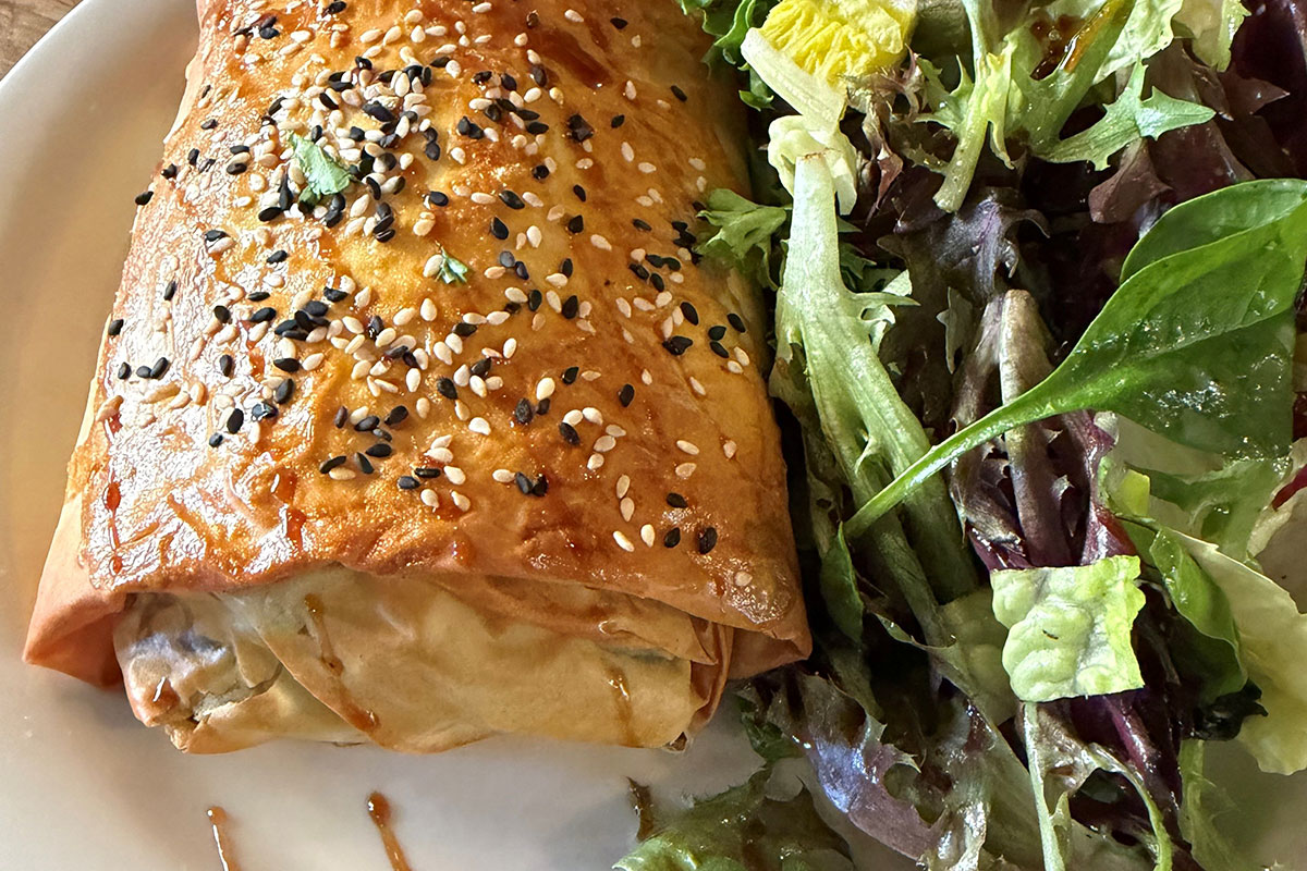 Close-up on a golden brown pastry topped with black and white sesame seeds alongside a green salad on a cardboard plate.