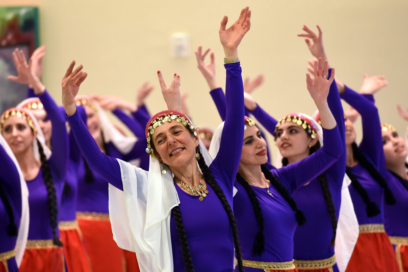 Women dancers in matching purple dresses of purple tops, red skirts, and gold belts, with white veils and long dark braids, dance with their hands raised above their heads.