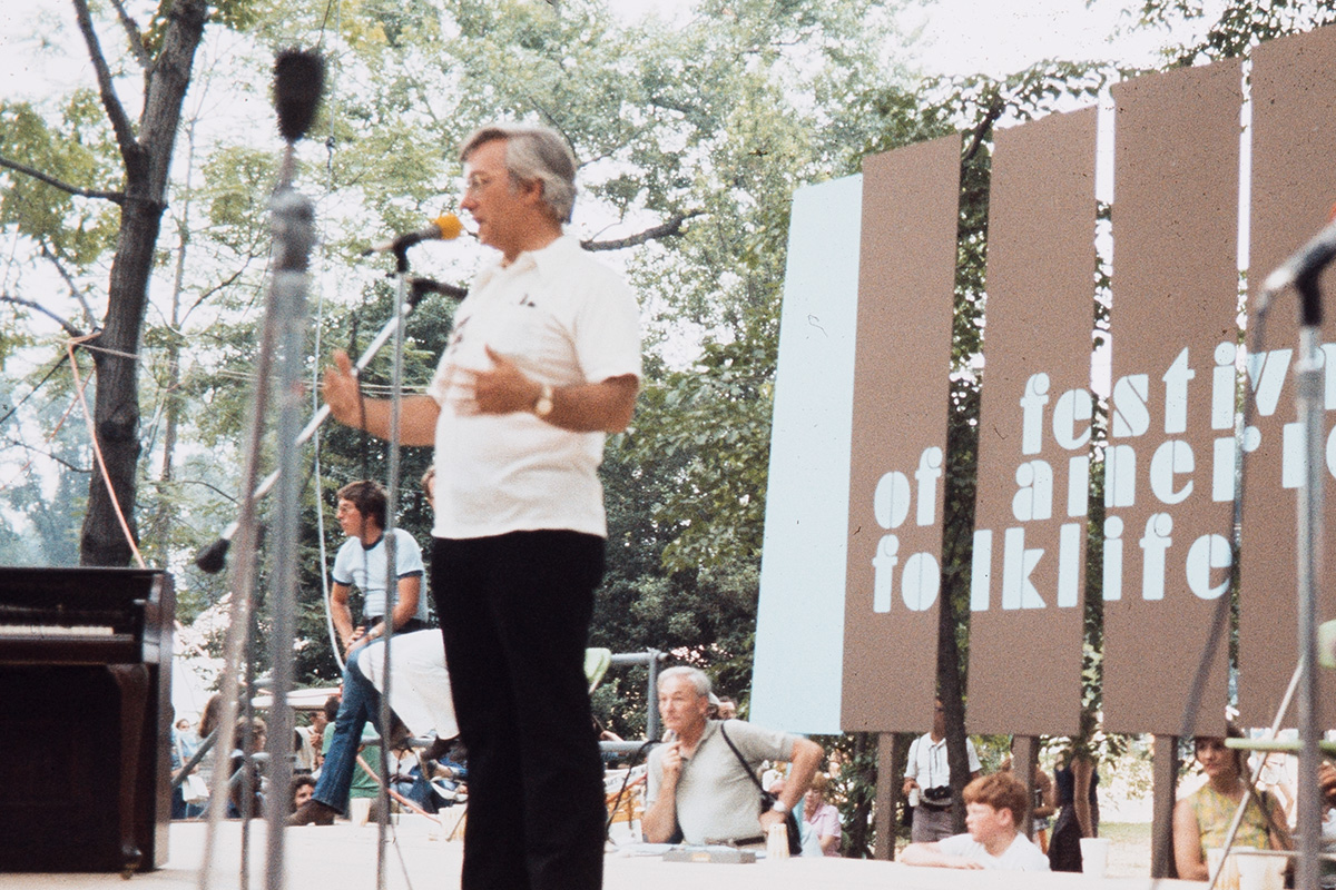 Old color photograph of a man standing at a microphone on stage, wearing a white dress shirt and black pants. Behind him, a stage backdrop with the words Festival of American Folklife, as well as people standing around and setting up equipment.