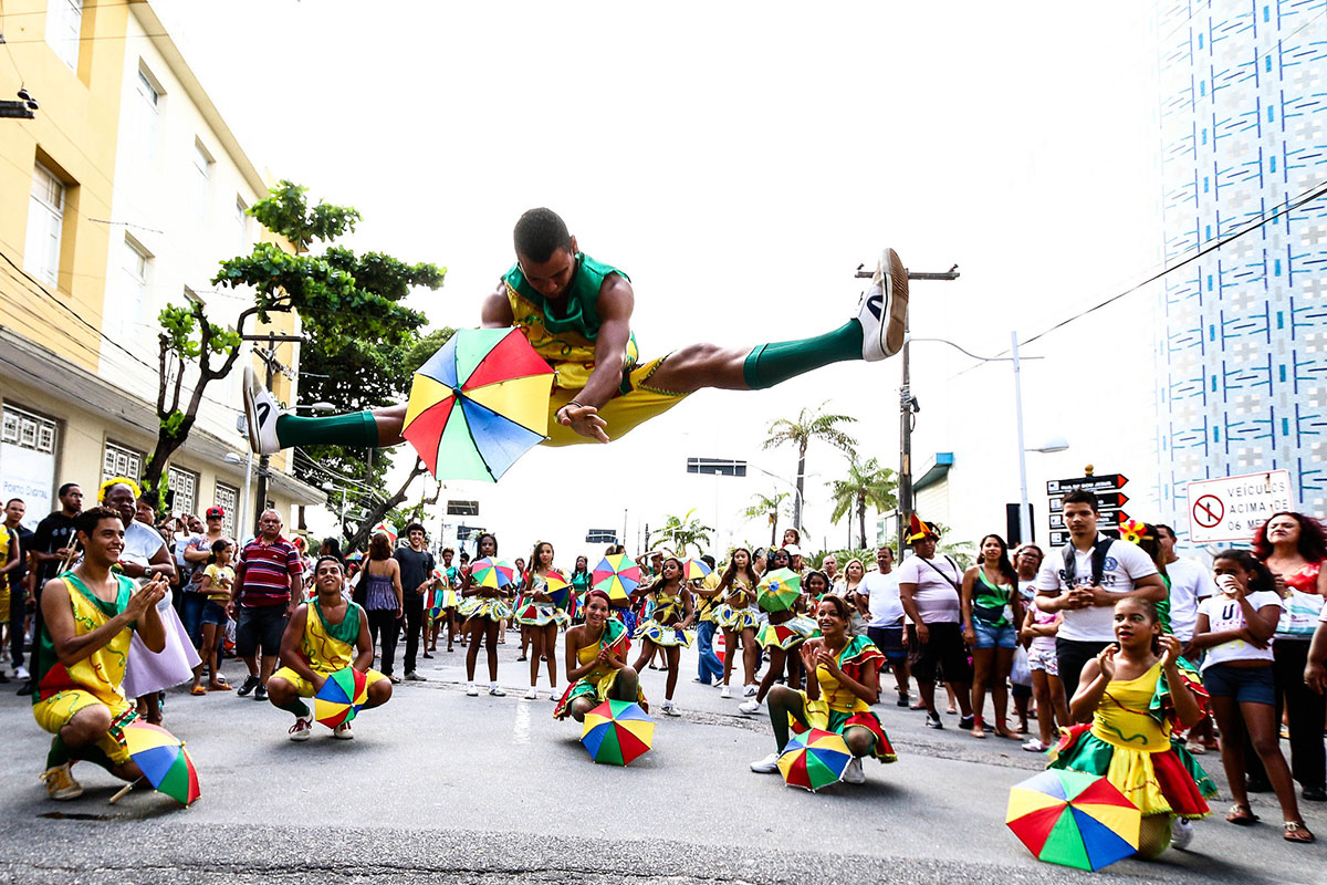 A dance troupe performs in a city street, wearing matching yellow and green uniforms. Five are knelt in a semi-circular around a sixth performer, maybe five feet in the air doing the splits. All hold small rainbow-colored umbrellas.