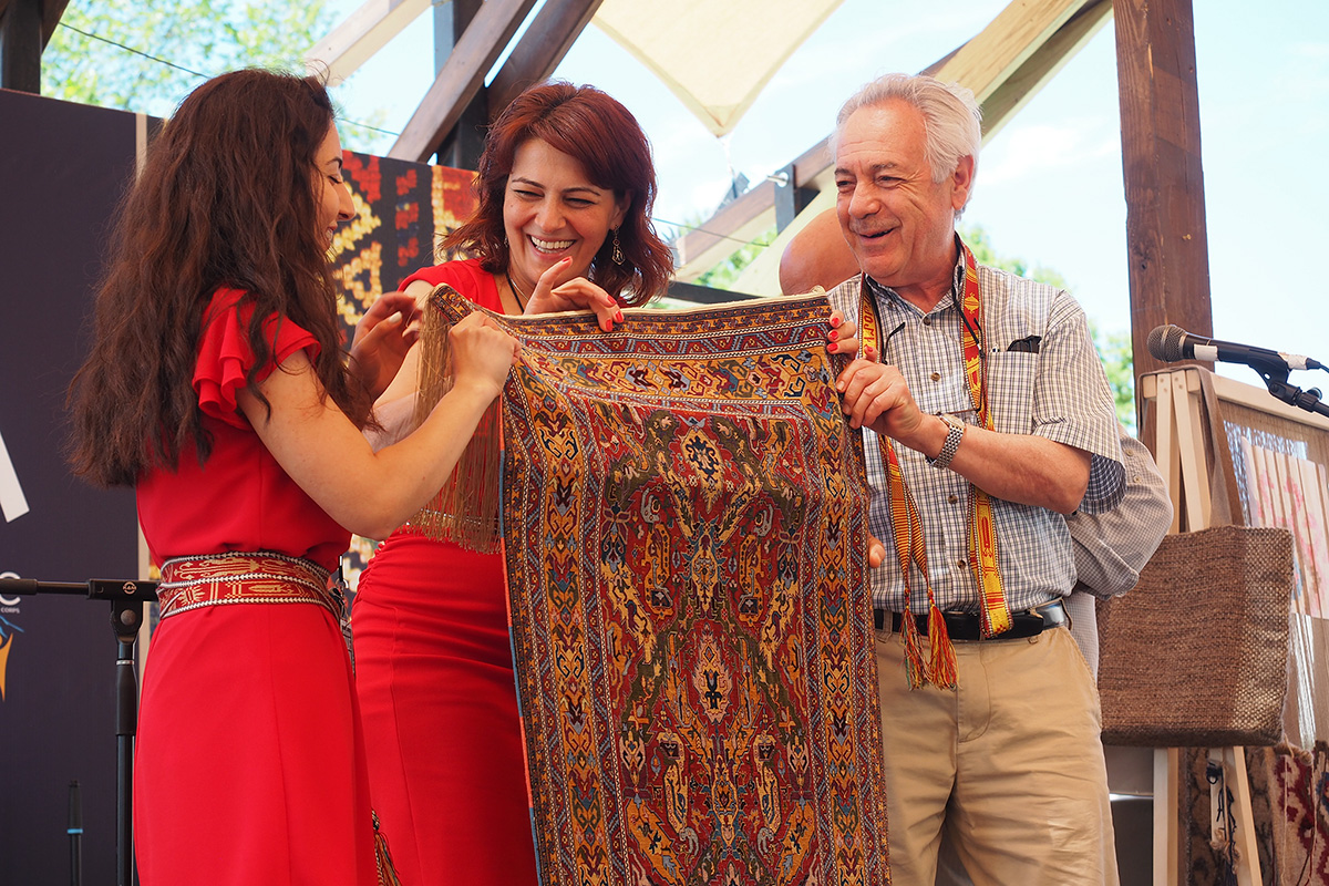 Weavers Diana Hovhannisyan and Ruzanna Torozyan along with presenter Levon Der Bedrossian show off the completed rug on the Hyurasenyak Stage at the 2018 Smithsonian Folklife Festival. 
