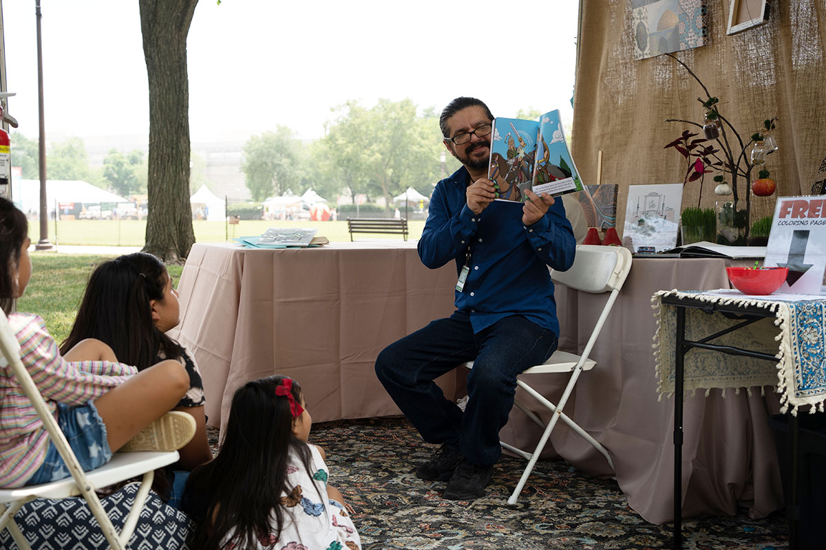 A man sitting in a folding chairs holds up an illustrated picture book, reading it to a group of kids seated on the left. Behind him, a table with ornaments hanging from a small display tree.