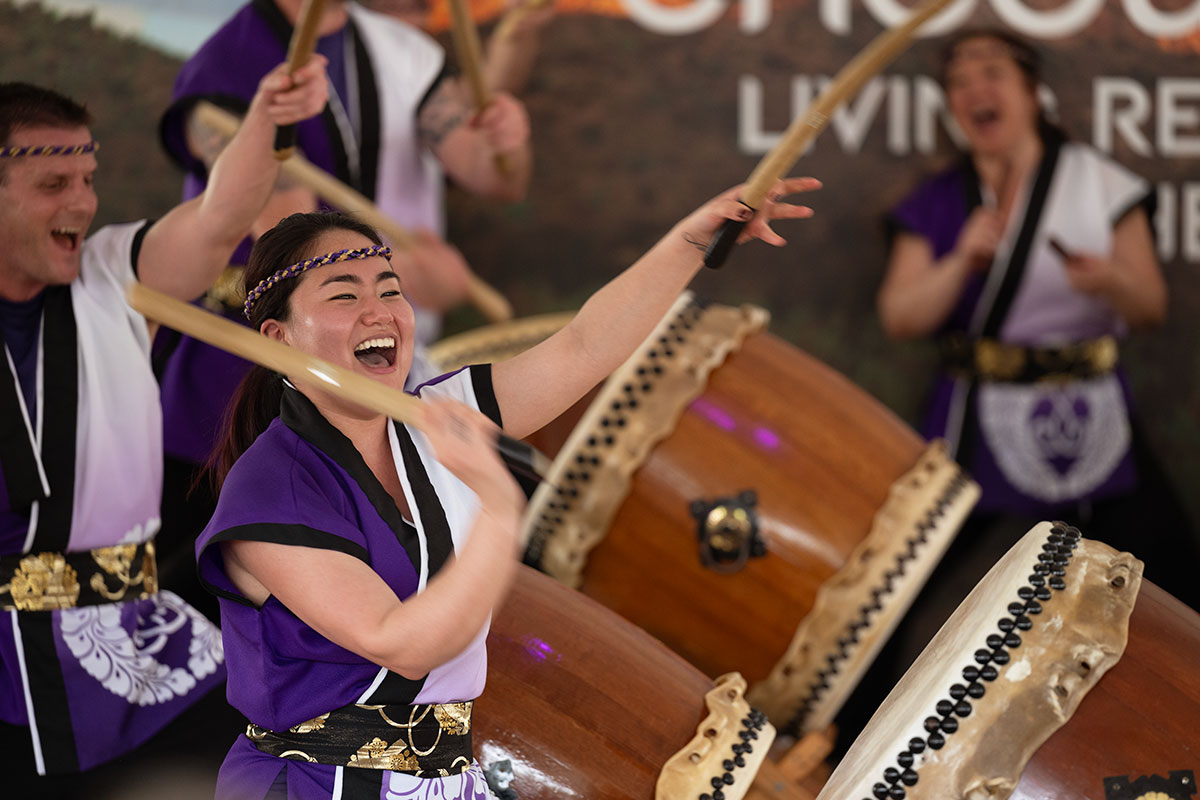 Four people beating wooden taiko drums with wooden sticks, each with their mouths wide open as if mid-yell.
