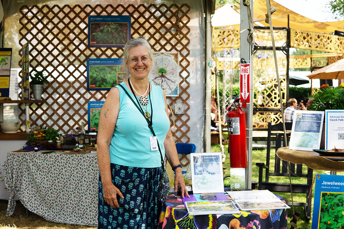 A woman in blue tank top and black and blue patterned skirt smiles under a tent decorated with plants and photos of plants.